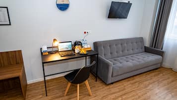 A modern living room with a grey sofa, a wooden floor, a desk with a laptop, a chair, a clock, and a TV mounted on the wall.