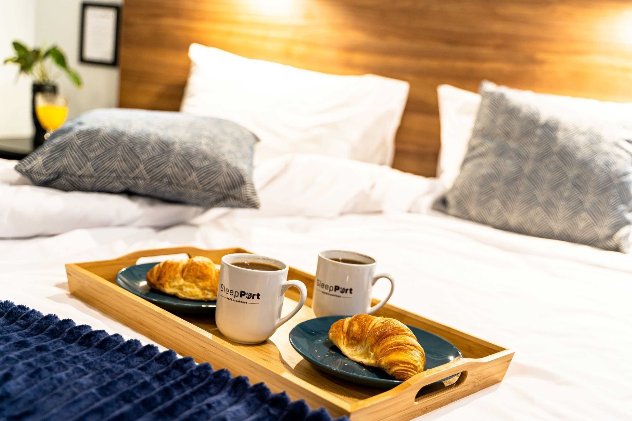 Un lit bien fait, avec une literie blanche et deux oreillers, un plateau avec des croissants et des tasses de café, et une plante en arrière-plan.