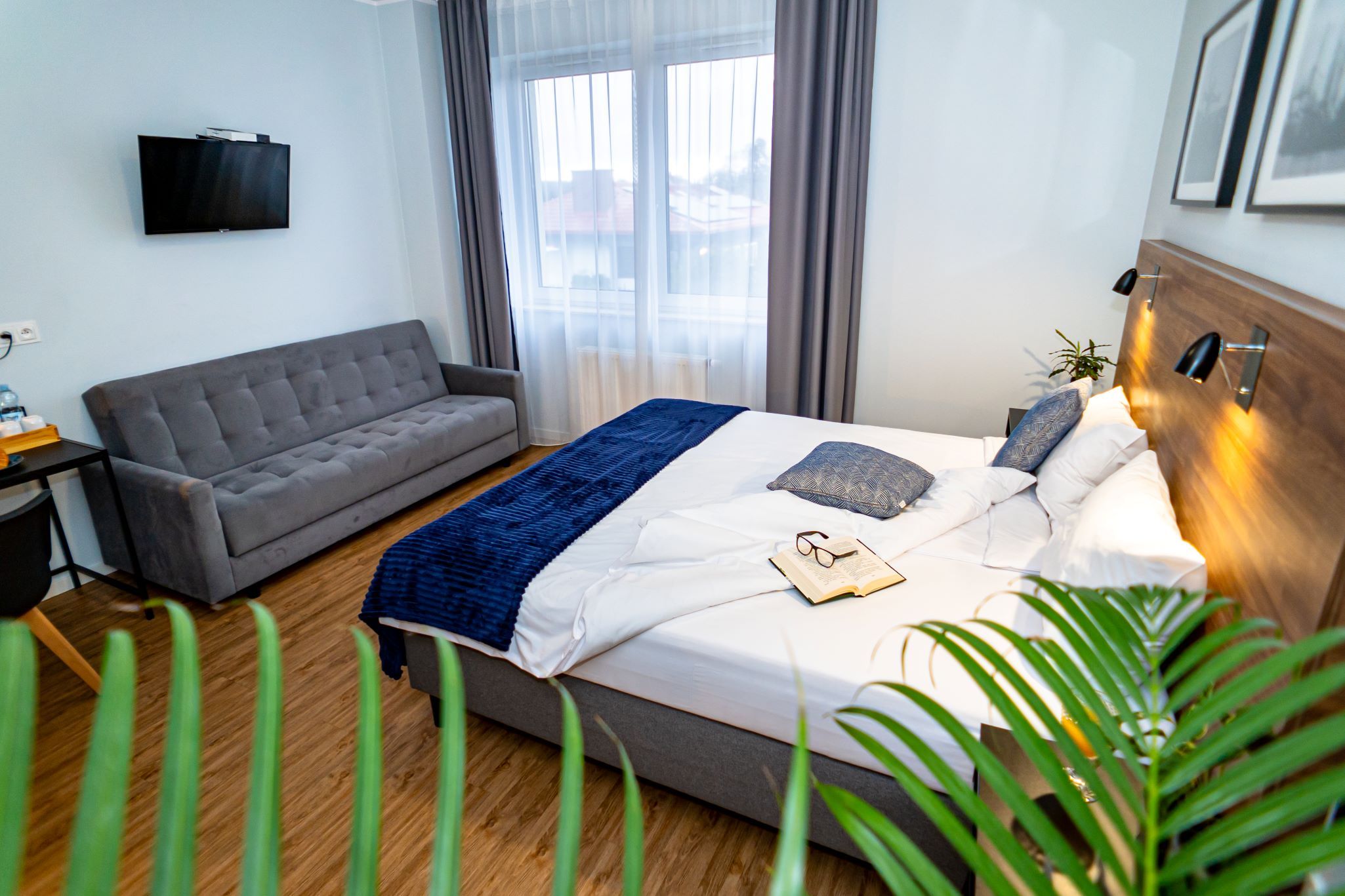 A modern hotel room with a bed, a grey sofa, a TV, and a window with curtains.
