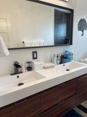 A bathroom with two sinks, a large mirror, and a wooden vanity with drawers. There are towels, toiletries, and a decorative wall piece.