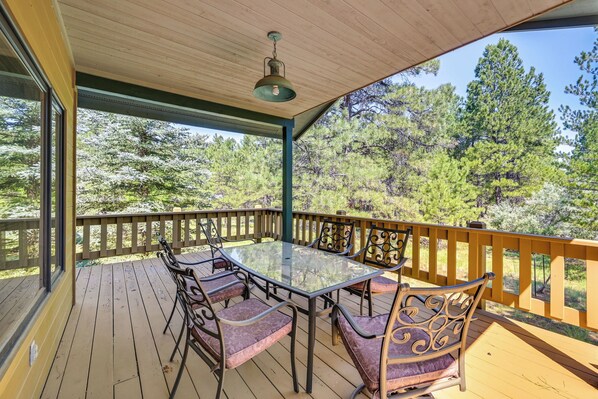 Terraza techada al aire libre con una mesa y sillas de vidrio, rodeada por una barandilla de madera, con vista a un área boscosa.