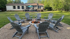 A circular outdoor seating area with Adirondack chairs around a fire pit, a covered patio with a grill, and a house in the background. - Revilo Timber Haven Mauston - Near The Dells and Castle Rock Lake! (Mauston)