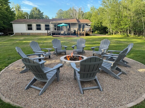 A circular outdoor seating area with Adirondack chairs around a fire pit, a covered patio with a grill, and a house in the background. - Revilo Timber Haven Mauston - Near The Dells and Castle Rock Lake! (Mauston)