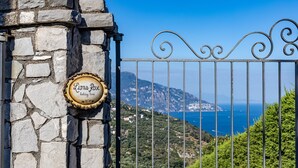 A stone wall with a sign reading "Lions Rose holiday home" and a metal gate with a scenic view of the sea and mountains in the background.