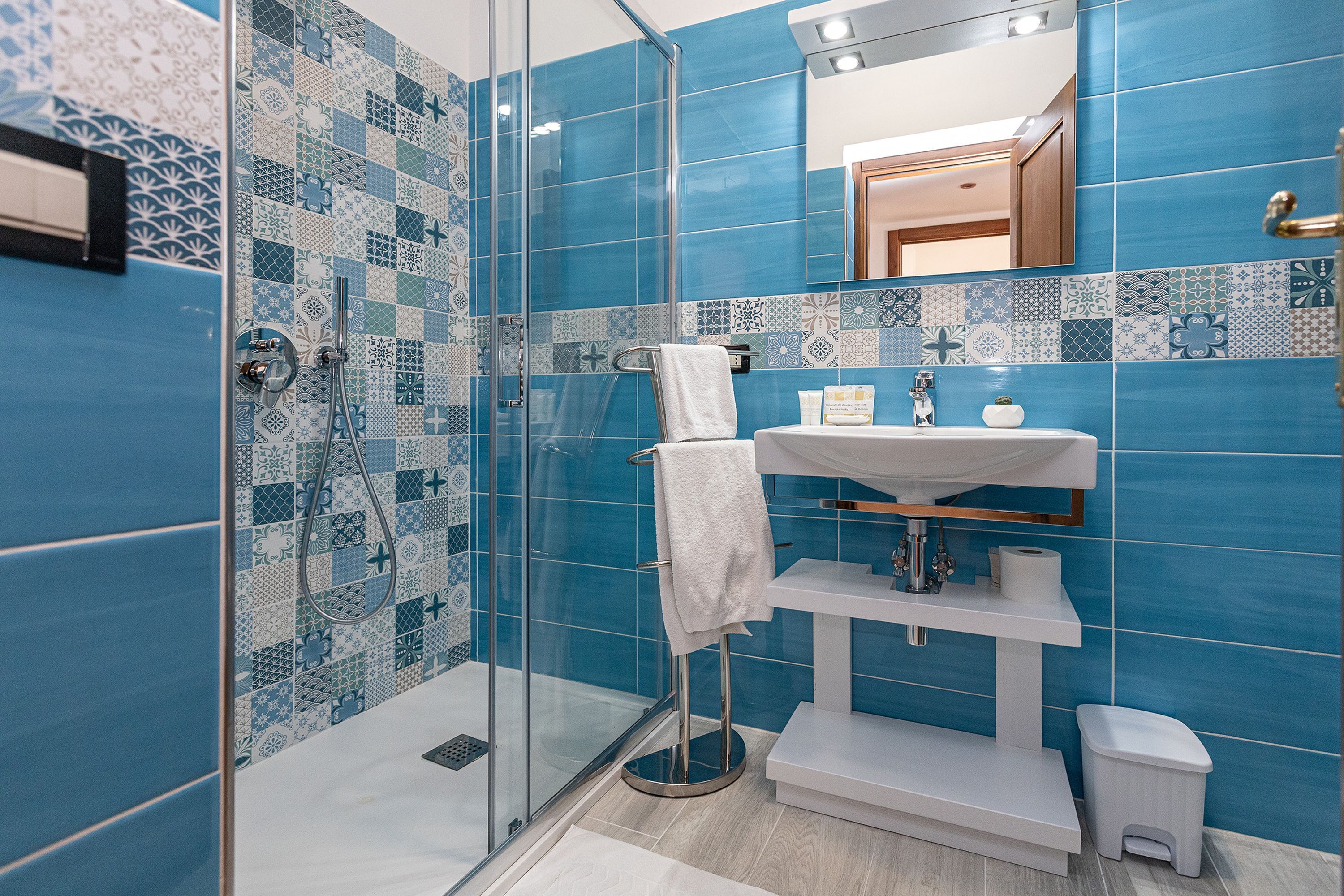 Une salle de bain avec des carreaux bleus à motifs, une cabine de douche en verre, un lavabo blanc et une petite poubelle blanche.