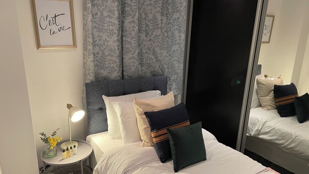 Una cama bien hecha con ropa de cama blanca y varias almohadas, una mesita de noche con una lámpara y un jarrón de flores, y un espejo grande que refleja la habitación.