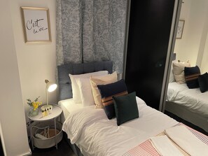 A neatly made bed with white bedding and multiple pillows, a bedside table with a lamp and a vase of flowers, and a large mirror reflecting the room. - Stepney Green Gated Area with Parking & Garden (London)