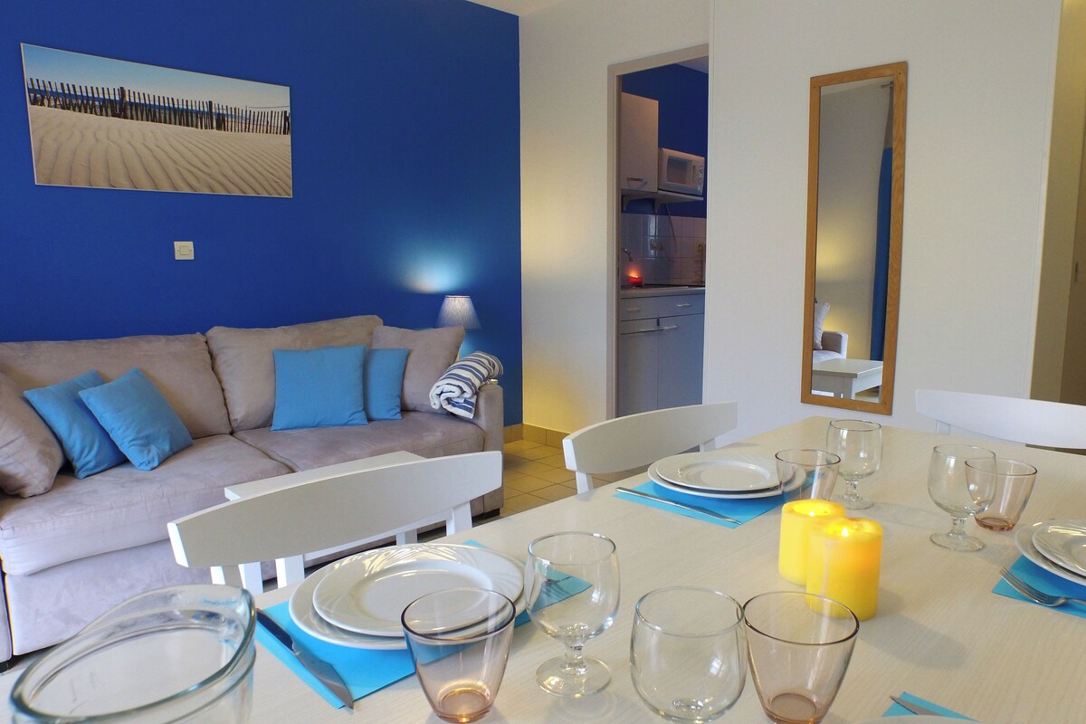 A dining area with a table set for four, a sofa with blue cushions, a kitchen in the background, and a large mirror on the wall.