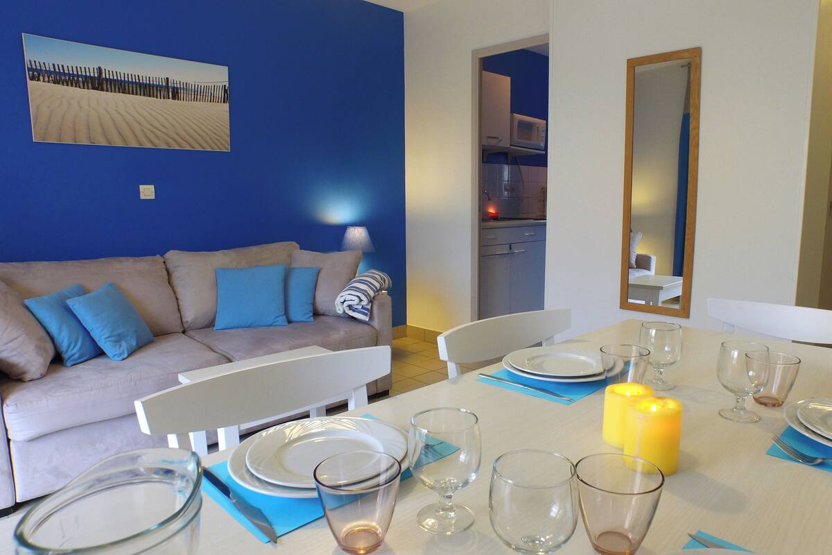 A dining area with a table set for four, a sofa with blue cushions, a kitchen in the background, and a large mirror on the wall.