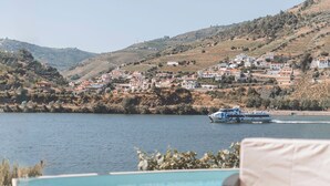 Área da piscina com vista para um lago e um barco. Há espreguiçadeiras e uma mesa com uma cesta de frutas.