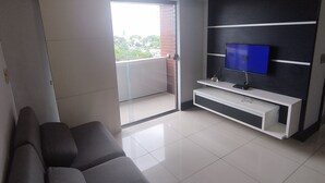 A modern living room with a grey sofa, a TV on a black and white entertainment unit, and a sliding glass door leading to a balcony. - Flat Adrianópolis All Suítes - Manaus (Manaus)