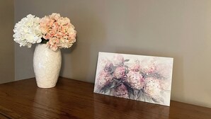 A wooden table with a white vase containing flowers and a framed picture of flowers. - Meadow Winds, A mountain getaway.  (Galax)