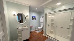 A bathroom with a wooden floor, a white sink with a mirror, a toilet, and a glass-enclosed shower. - Meadow Winds, A mountain getaway.  (Galax)