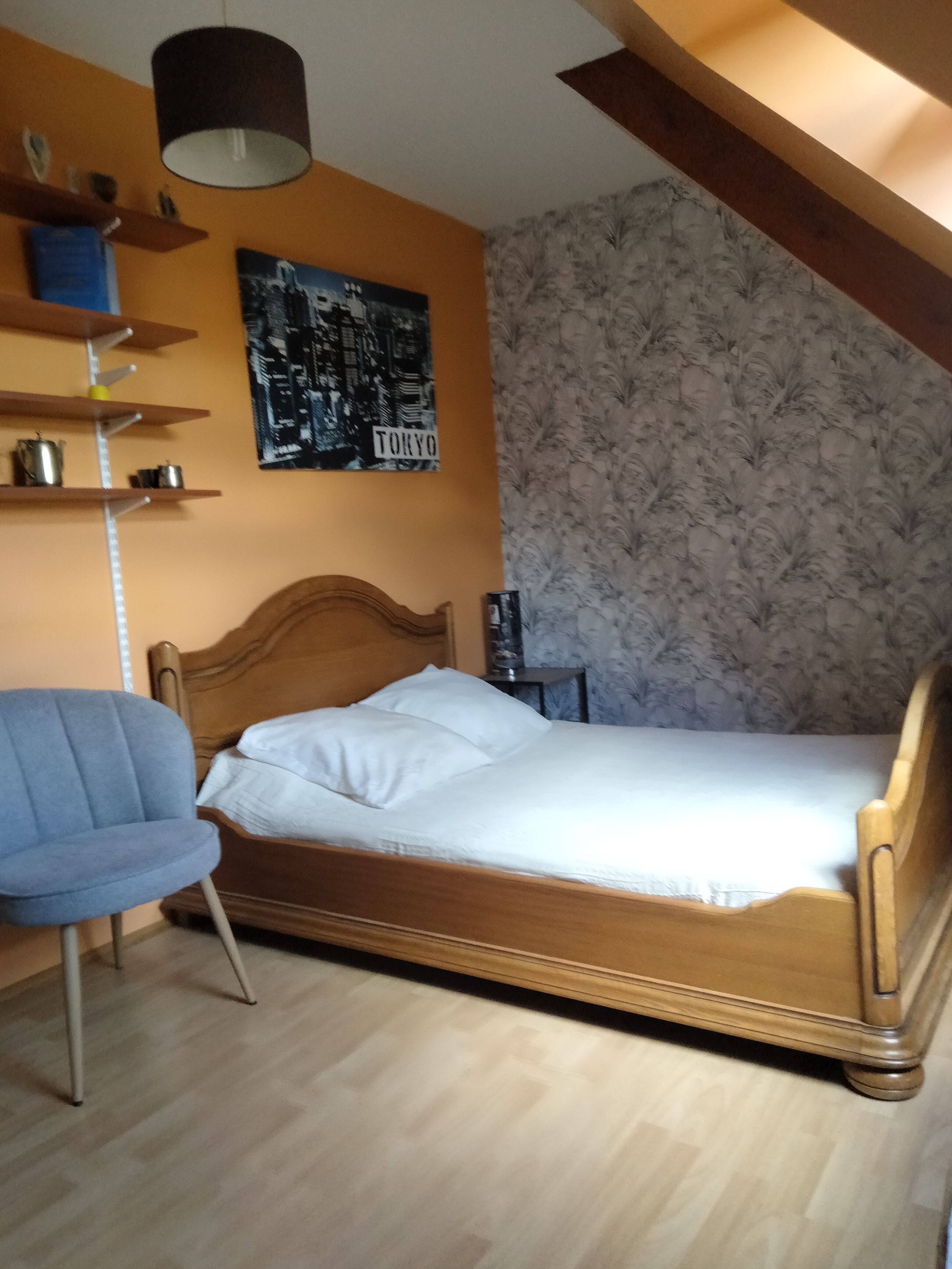 A bedroom with a wooden bed, a blue chair, a shelf with items, a poster on the wall, and a patterned wallpaper.