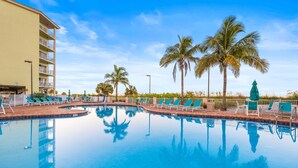 A swimming pool with lounge chairs and palm trees, surrounded by a building with balconies. - Beachfront Escape: 2BR Condo, Tiki Bar (Treasure Island)