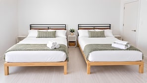 Two twin beds with wooden frames and white bedding, a small bedside table with a plant, and a white door in a minimalist room. - Suite Life Wilmington-Spacious, Pet-Friendly 1BR, Pool, Balcony, Prime Location (Wilmington)