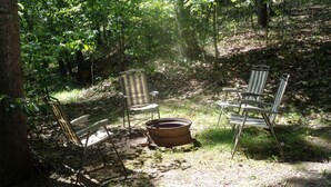 A rustic outdoor seating area with metal chairs and a large metal pot, surrounded by trees and natural ground cover. - The Cottage in the Woods, near Deersville and Tappan and Clendening Lakes (Deersville)