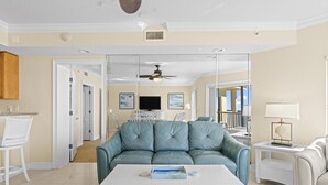 A spacious living room with a light blue sofa, a white coffee table, and a white armchair. There is a television mounted on the wall and a ceiling fan above. - 3/3 Penthouse Condo with Epic Beach Views (Treasure Island)