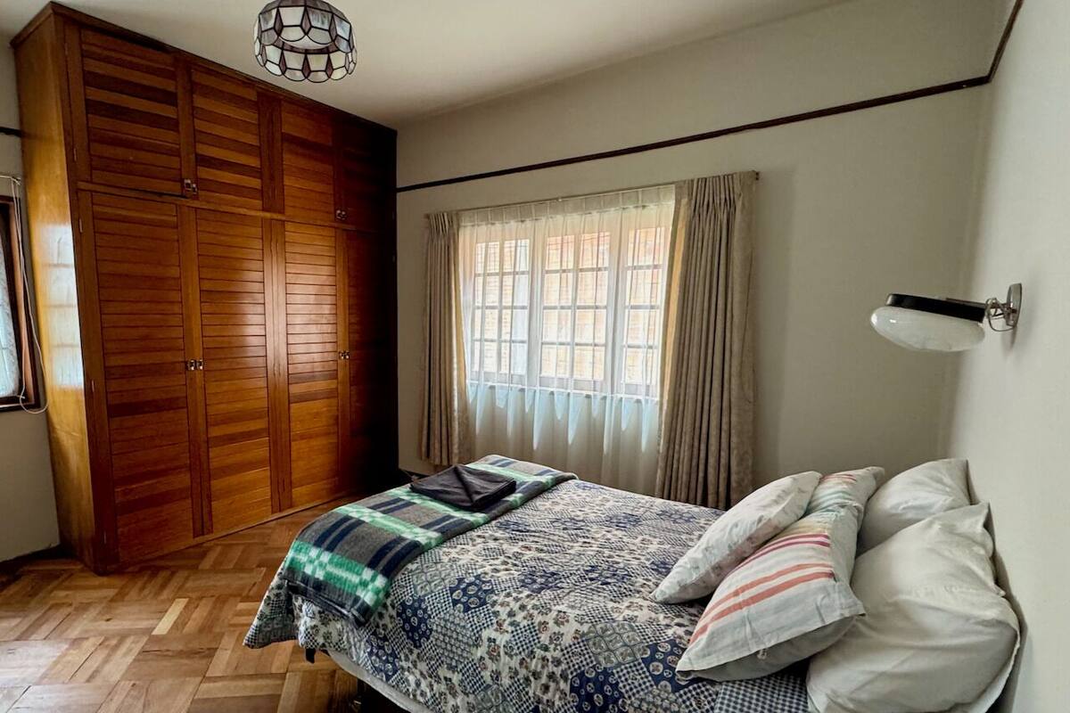 A bedroom with a wooden wardrobe, a bed with patterned bedding, a window with curtains, and a wall-mounted light fixture.
