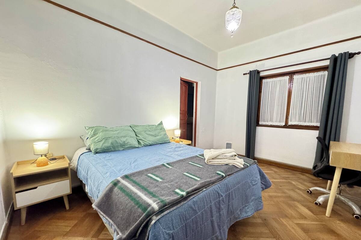 A neatly arranged bedroom with a bed, bedside tables, a desk, and a window with curtains.