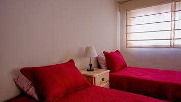 Quarto com duas camas, cada uma com roupa de cama vermelha, uma mesa de cabeceira com abajur e uma janela com persianas.