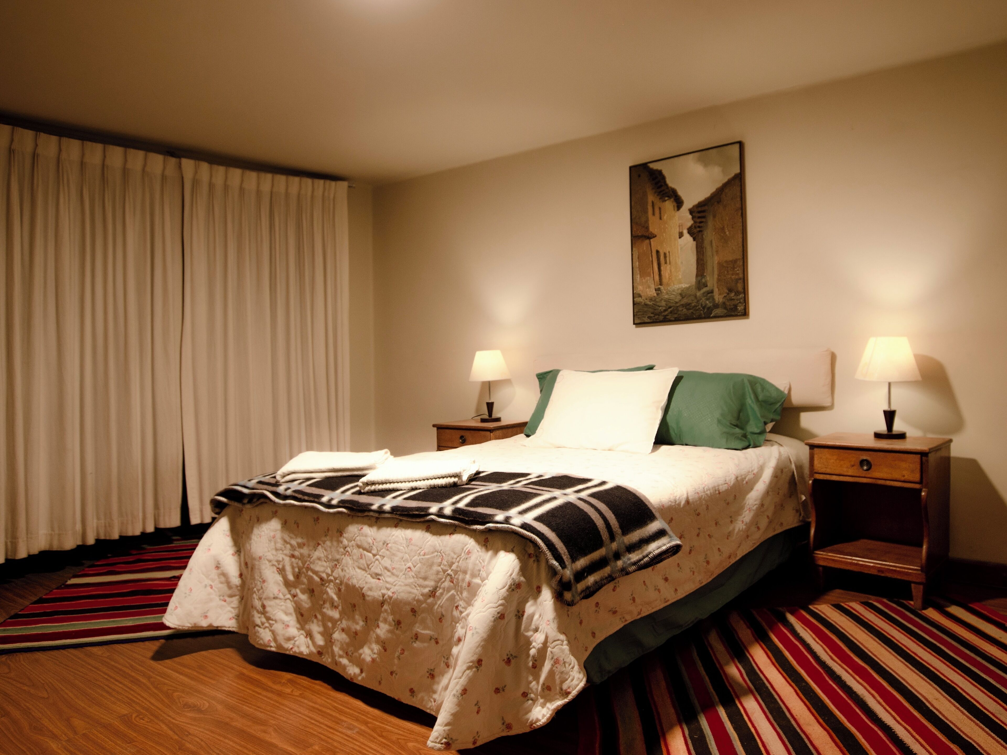 Een hotelkamer met een bed, twee nachtkastjes met lampen, een schilderij aan de muur en een gestreept tapijt.