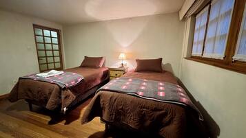 Quarto com duas camas de solteiro, mesa de cabeceira com luminária, piso de madeira e janela com cortinas.