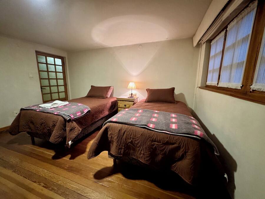 Quarto com duas camas de solteiro, mesa de cabeceira com luminária, piso de madeira e janela com cortinas.