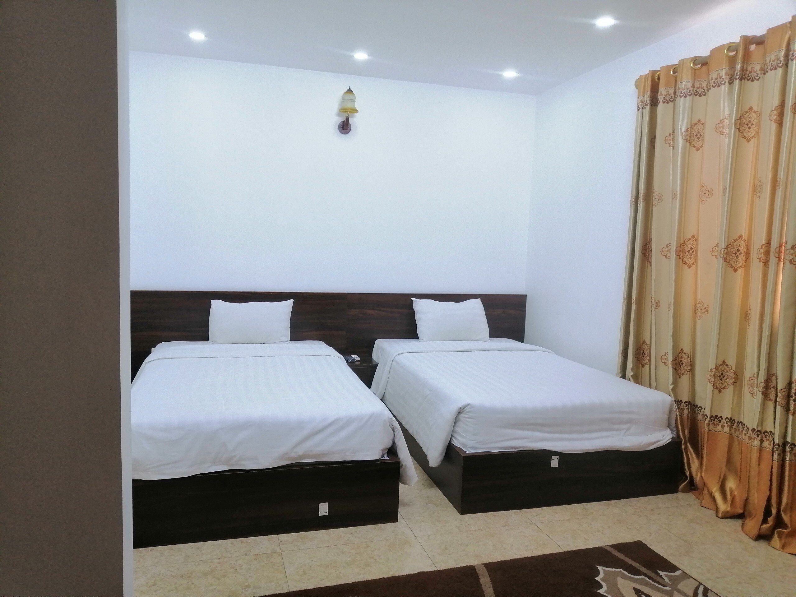Habitación de hotel con dos camas individuales, cabecero de madera, luz de pared y cortina estampada.