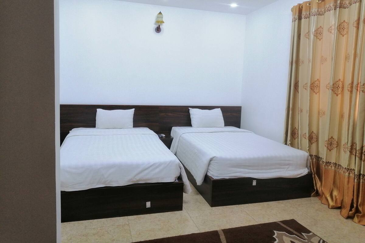 A hotel room with two single beds, a wooden headboard, a wall-mounted light, and a patterned curtain.