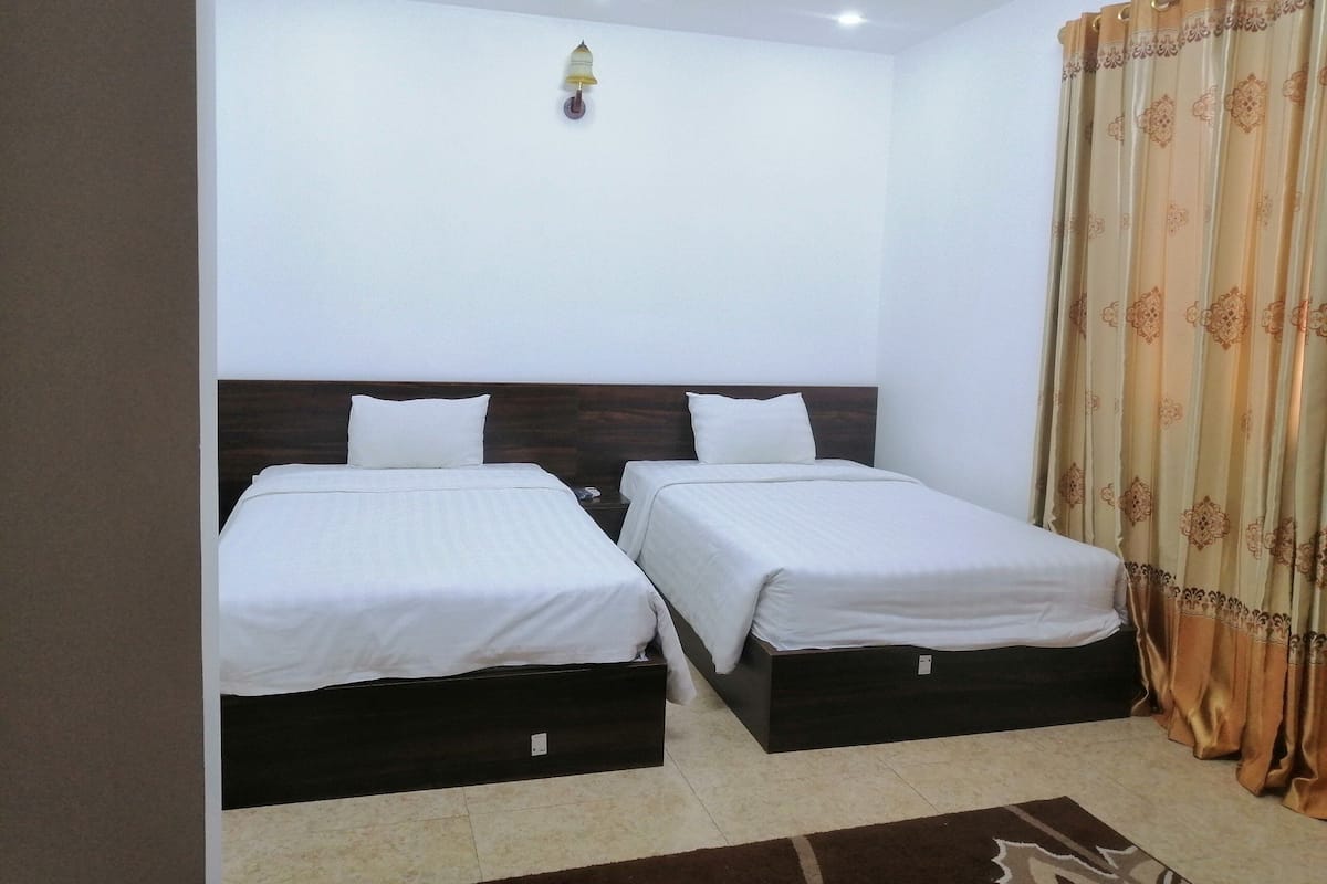 A hotel room with two single beds, a wooden headboard, a wall-mounted light, and a patterned curtain.
