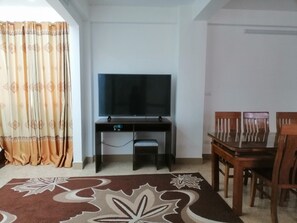 Una habitación con televisión sobre un mueble, una mesa de comedor con sillas y una alfombra estampada.