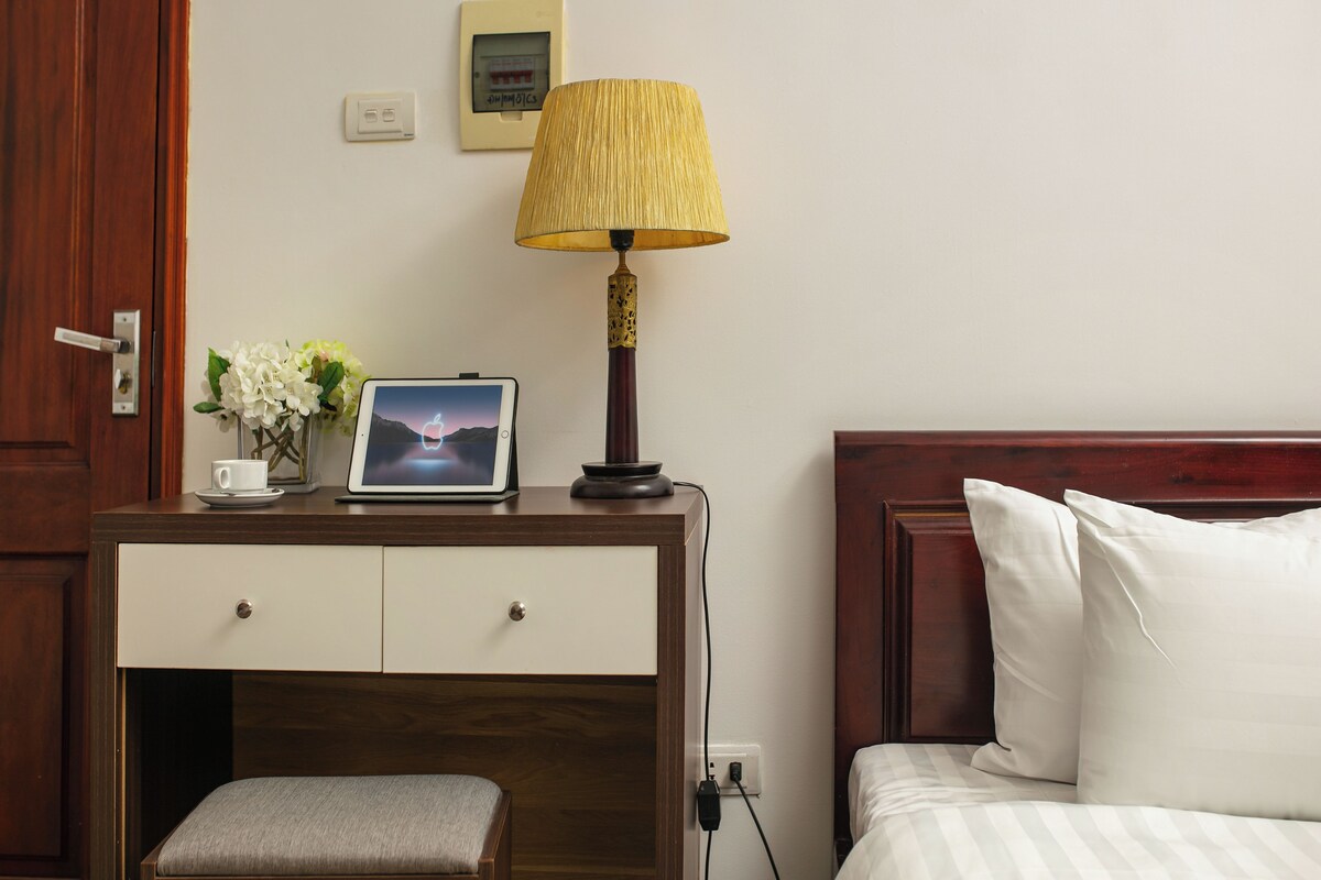 Habitación de hotel con escritorio de madera, lámpara, cuadro enmarco, pequeño jarrón con flores y cama con almohadas blancas.