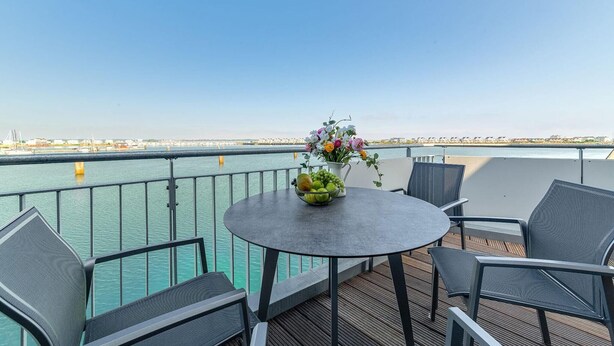 Eine Terrasse mit einem runden Tisch, Stühlen, einer Blumenvase und Blick auf das Wasser sowie entfernte Gebäude.