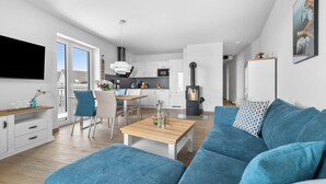 Eine moderne Wohnung mit Wohnzimmer, Essbereich und Küche. An der Wand ist ein großer Flachbildfernseher angebracht, ein Sofa mit gemusterten Kissen und ein Couchtisch aus Holz.