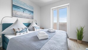 Ein modernes Schlafzimmer mit einem großen Bett, zwei Kissen, einem Nachttisch, einem Bild mit Strandmotiv und Meerblick durch eine Schiebetür.