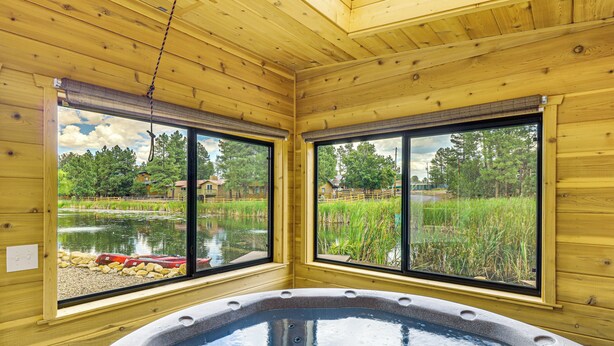 Una cabaña de madera con jacuzzi, ventanas amplias y vistas a un estanque y follaje.