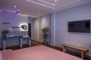 Un dormitorio moderno con una cama, un televisor montado en la pared, una mesita de noche, un banco, un lavabo con espejo y una planta en maceta.