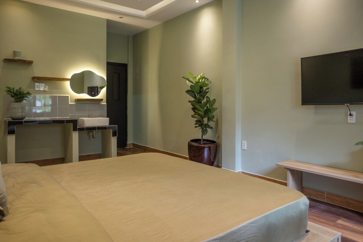 Habitación de hotel con una cama grande, un escritorio con silla, un lavamanos, un espejo, una planta en maceta y un televisor montado en la pared.