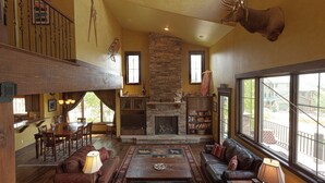 A spacious living room with a stone fireplace, leather sofas, a dining area, and large windows offering a view of the outdoors.