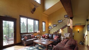 A spacious living room with a large window, a fireplace, a staircase, and a mounted deer head.