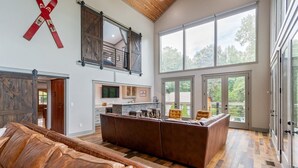 A spacious living area with a large leather couch, a wooden coffee table, and a kitchen area with a large island. There are large windows providing natural light and views of the outdoors.