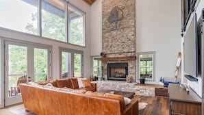 A spacious living room with a large leather sofa, a stone fireplace, and large windows offering a view of the outdoors.