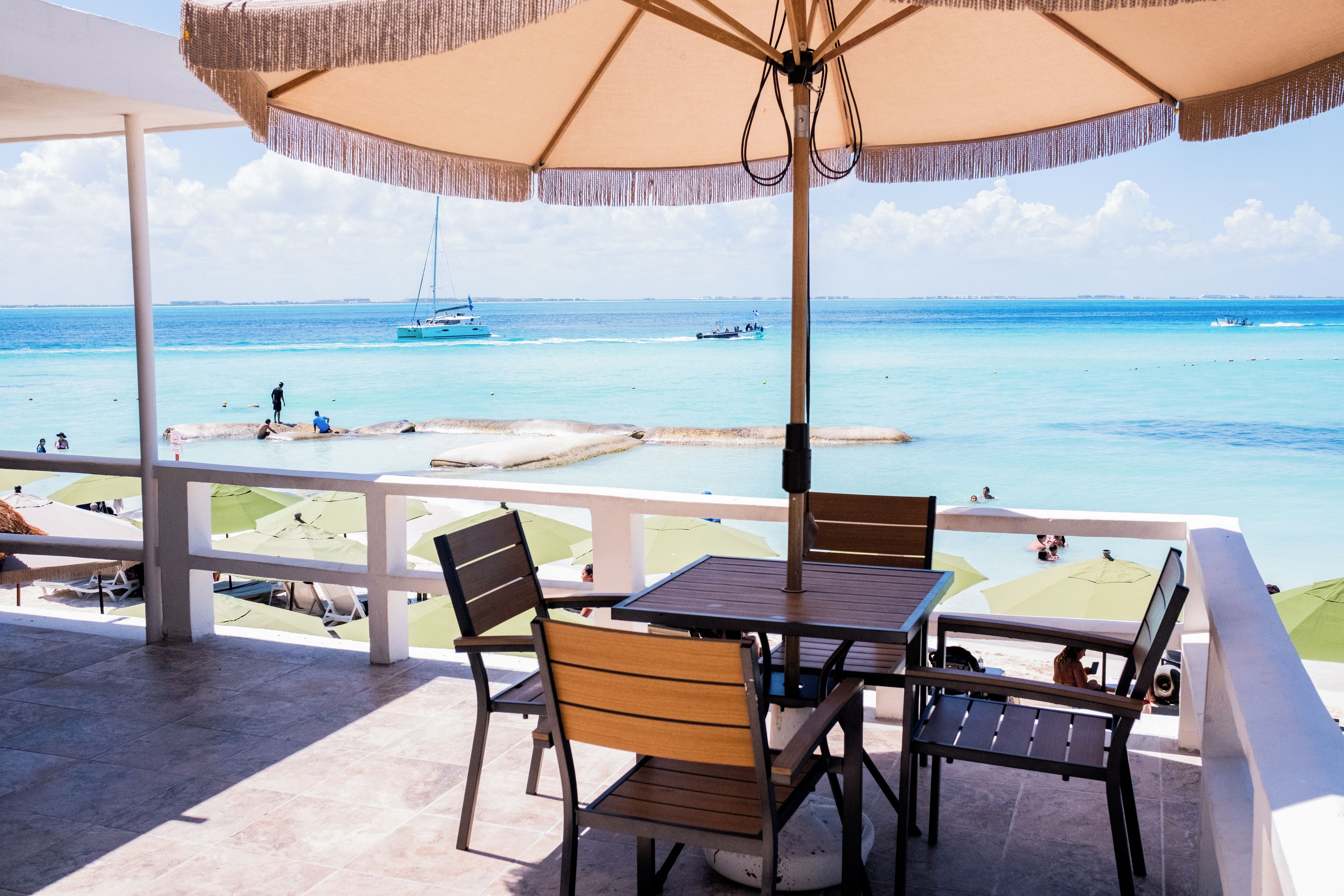 A terrace with a table and chairs, overlooking a beach with boats and people.