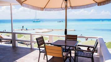 A terrace with a table and chairs, overlooking a beach with boats and people.