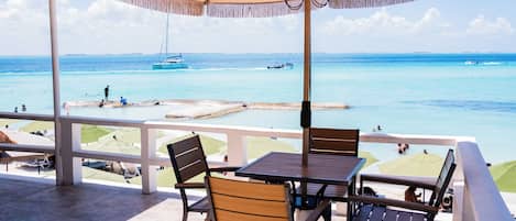 A terrace with a table and chairs, overlooking a beach with boats and people.
