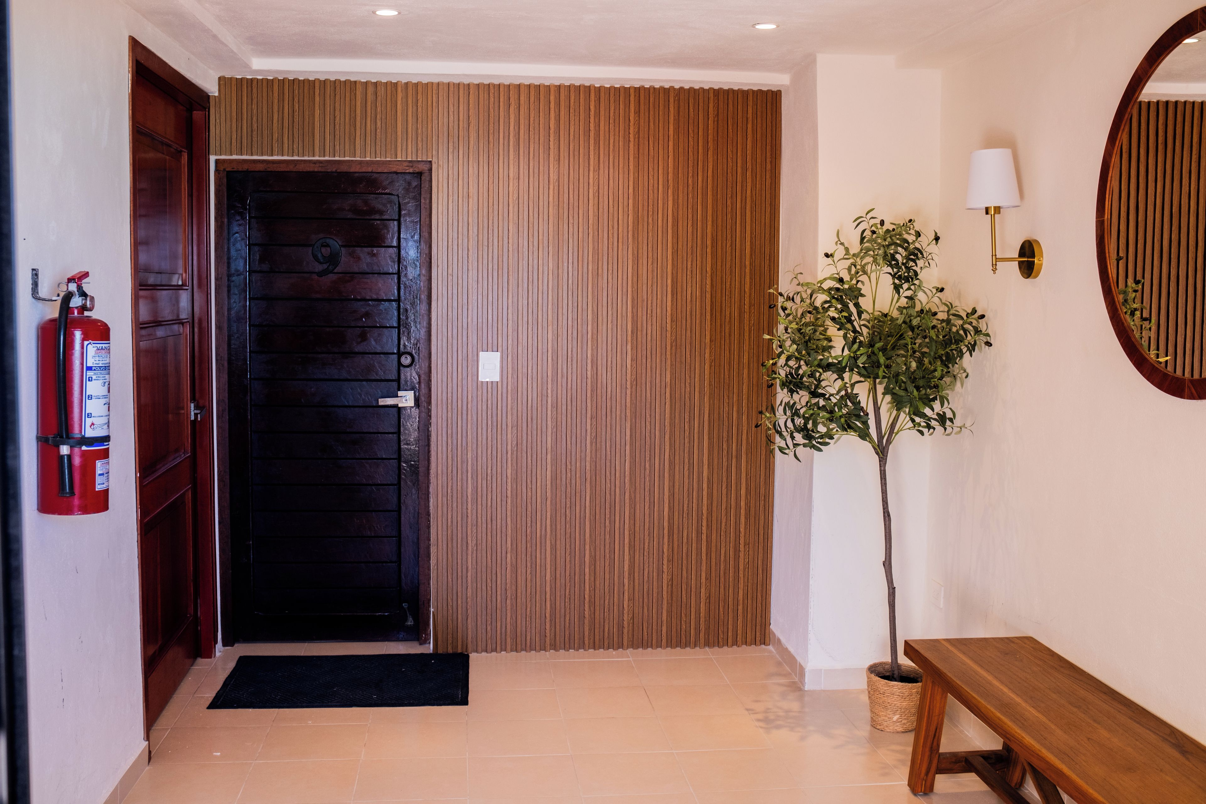 A wooden door, a fire extinguisher, a potted plant, a bench, and a wall-mounted lamp.