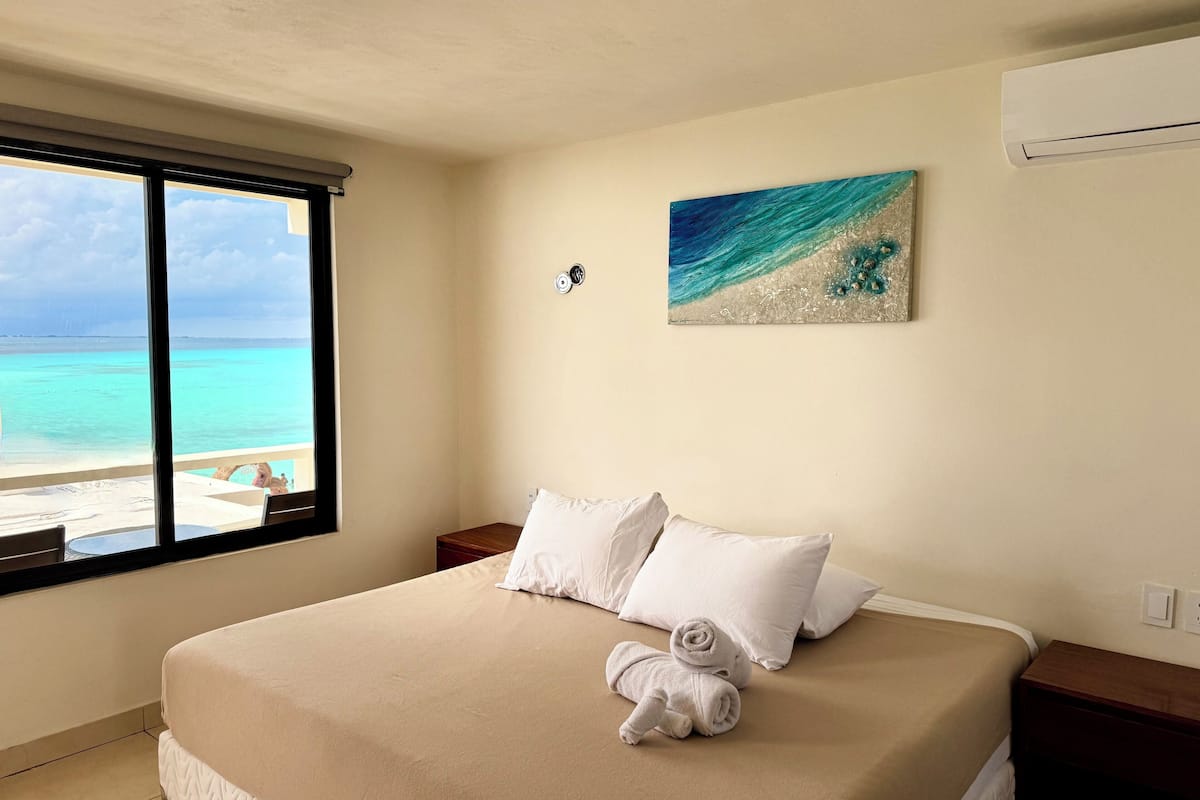 A hotel room with a bed, a painting of a beach scene, and a view of the ocean through a large window.