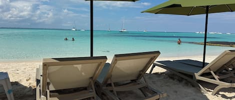 Une plage avec des chaises longues et des parasols, une vue dégagée sur l’océan et des voiliers à l’horizon.