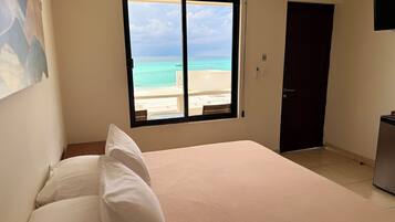 A hotel room with a bed, a window with a view of the beach, a door, and a small table.